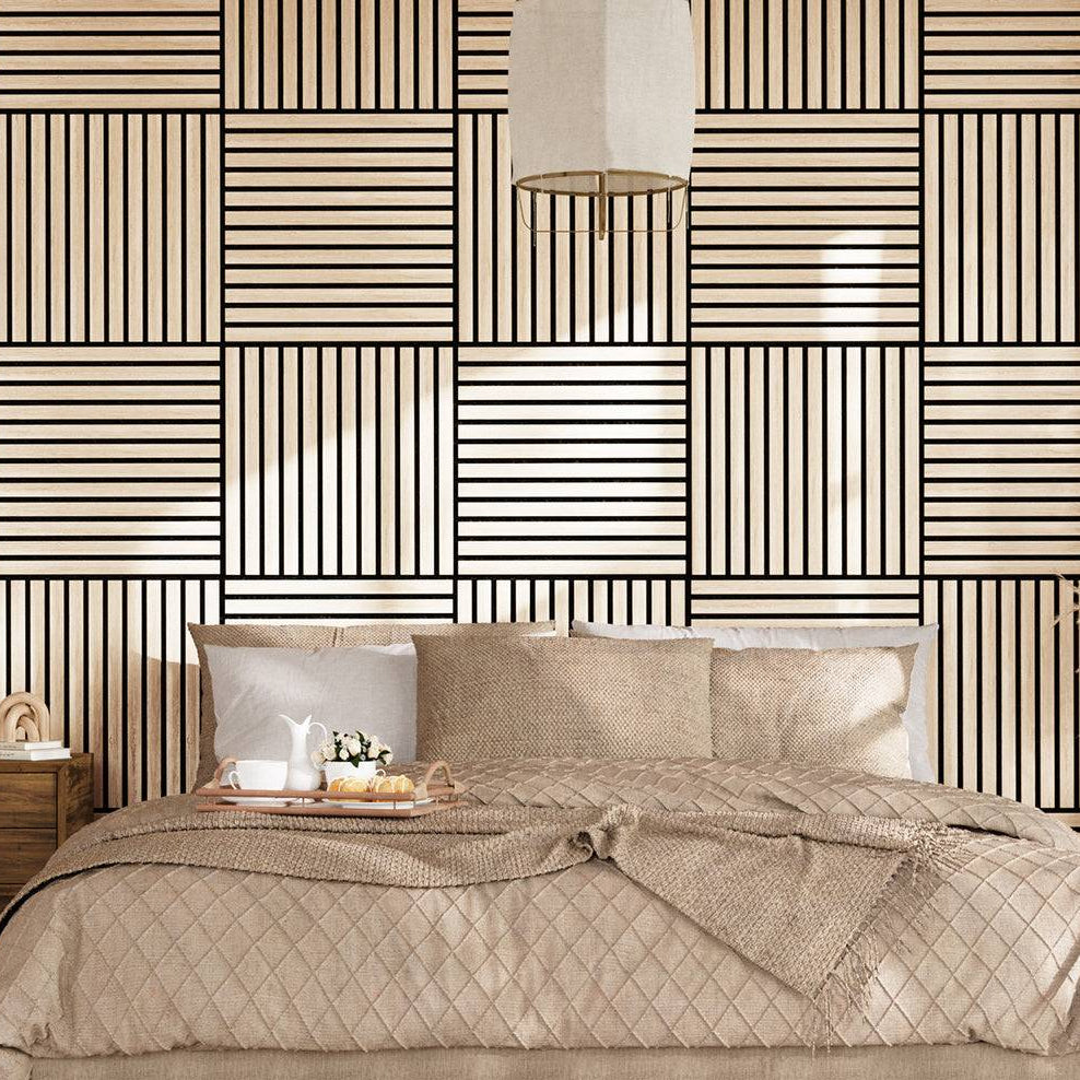 A styled bedroom scene featuring a bed dressed in beige quilted bedding and pillows, set against a bold geometric wallpaper with black and beige stripes. A matching patterned rug covers the floor, and a wooden nightstand with a lamp is visible on the left.