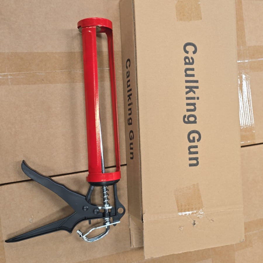 A red and black caulking gun resting next to its brown cardboard packaging box on a floor covered with protective plastic wrap.